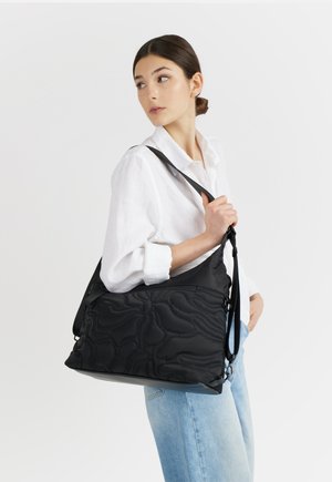 Giovane donna con camicia bianca e jeans blu che porta una grande borsa a tracolla nera trapuntata con tracolla regolabile, guardando di lato.