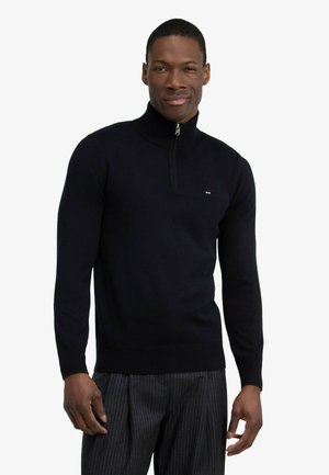 Homme portant un pull noir à manches longues avec un col à fermeture éclair un quart et un pantalon sombre à fines rayures, debout devant un fond uni.