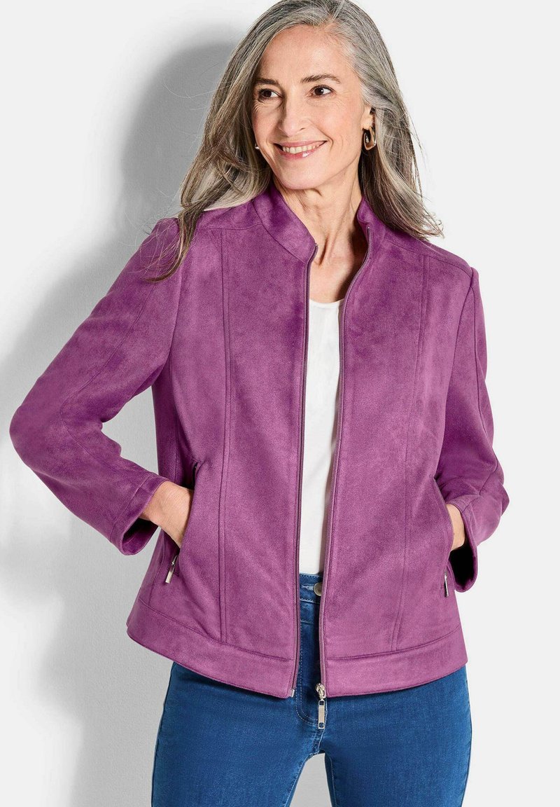 Veste en suède violet avec un col montant et une fermeture éclair. Comprend des poches latérales et des détails de couture, associée à un haut blanc et un jean bleu.