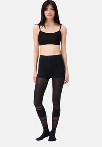 Schwarzes Bralette mit dünnen Trägern und Logo-Band, kombiniert mit hochgeschnittenen schwarzen Leggings, die transparente Einsätze und durchgehende Streifen an den Beinen aufweisen.