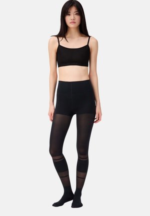 Schwarzes Bralette mit dünnen Trägern und Logo-Band, kombiniert mit hochgeschnittenen schwarzen Leggings, die transparente Einsätze und durchgehende Streifen an den Beinen aufweisen.