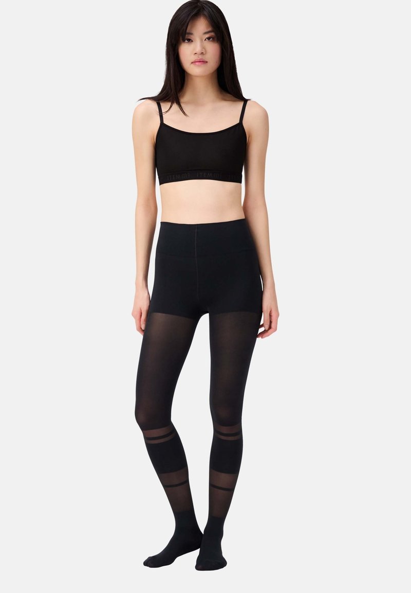 Schwarzes Bralette mit dünnen Trägern und Logo-Band, kombiniert mit hochgeschnittenen schwarzen Leggings, die transparente Einsätze und durchgehende Streifen an den Beinen aufweisen.