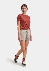 Roestkleurig T-shirt met korte mouwen, gecombineerd met lichtgrijze broekjes met trekkoord, en zwarte sneakers met witte zolen, voorzien van rood en blauw gestreepte sokken.