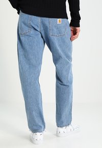 Osoba nosząca jasnoniebieskie dżinsy prostym kroju i białe trampki z czarnymi falistymi wzorami, stojąca na tle białego tła.