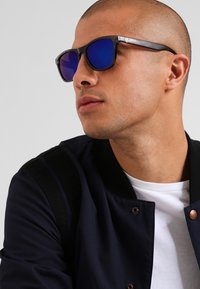 Ochelari de soare negri cu lentile albastre reflectorizante și un design elegant, unghiular. Purtați cu o jachetă bleumarin cu o textură și detalii subtile.