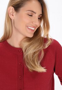 Sarkanais adītais kardigans ar apaļu kakla izgriezumu, piecu pogu aizdari un ribota apdare uz apkakles un pogu cilpām. Gluda tekstūra.