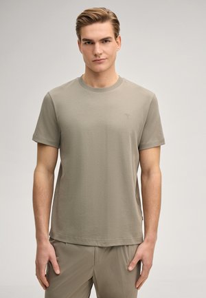 Joven con cabello corto, vistiendo una camiseta lisa color topo y pantalones a juego, de pie contra un fondo neutro claro.