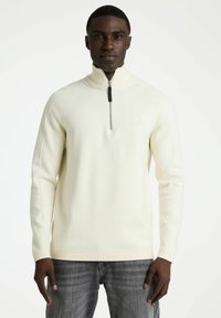 Jonge man in een crèmekleurige ritstrui met hoge kraag en vervaagde grijze jeans, staand tegen een effen witte achtergrond.