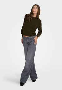 Femme portant un pull vert foncé et un jean gris à jambes larges, debout les jambes croisées, une main posée sur la hanche, regardant sur le côté.