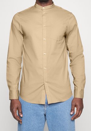 Homme portant une chemise beige à manches longues avec col mandarin et un jean bleu clair, debout devant un fond uni.