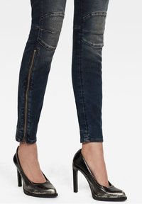 Vaqueros de mezclilla oscura con zonas desgastadas y costuras en panel, combinados con zapatos de tacón alto metálicos negros que tienen un corte en la punta y acentos texturizados.