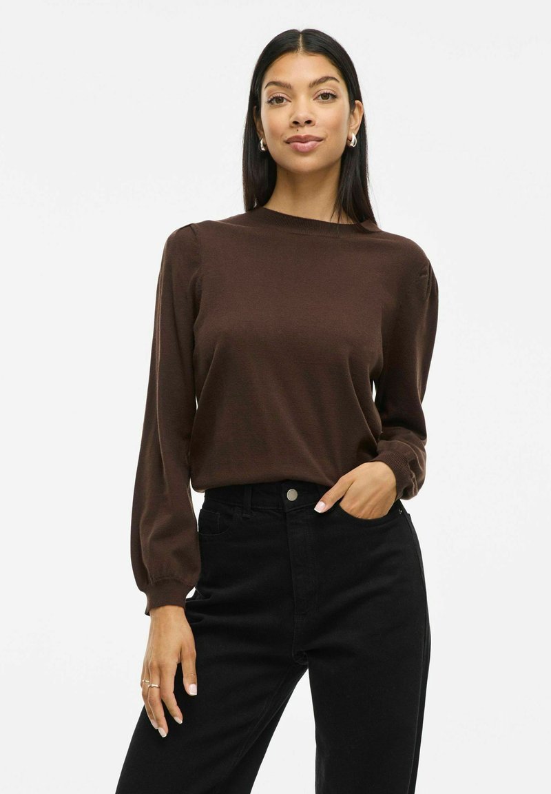 Top marrone a maniche lunghe con scollo rotondo e dettaglio delle spalle a palloncino, abbinato a jeans neri a vita alta. Tessuto liscio, vestibilità casual.
