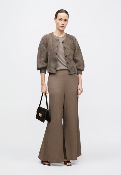 Femeie purtând pantaloni largi taupe, top asortat, cardigan scurt pufos, ținând o geantă de mână neagră, stând în fața unui fundal simplu și deschis la culoare.