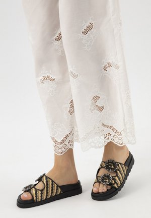 Pieds portant des sandales slides tissées noires et beiges avec des boucles décoratives, associées à un pantalon blanc en dentelle brodée.