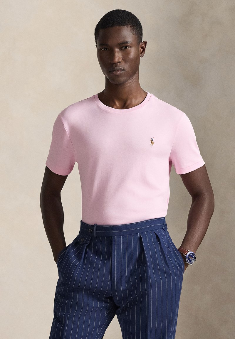 Šviesiai rožinė medvilninė marškinėliai su apvalia iškirpte ir trumpomis rankovėmis, ant krūtinės mažas logotipas, derinami su tamsiai mėlynomis dryžuotomis kelnėmis.