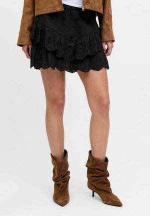 Femme portant une mini-jupe noire à volants avec des détails œillets, des bottines en daim marron décontractées et une veste courte marron.