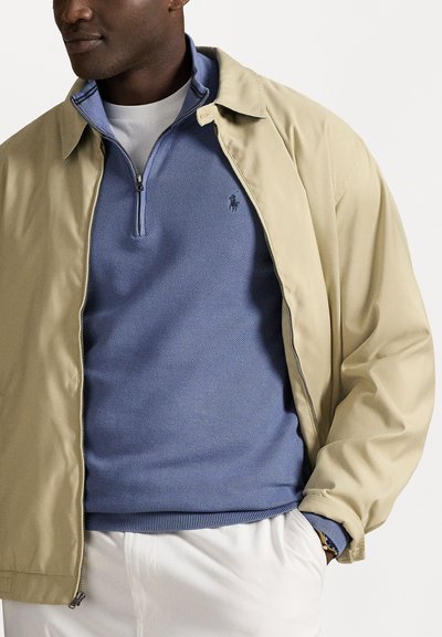 Beige zip-up jack over een lichtblauwe textuurtrui, gecombineerd met witte pants. Bevat een kraag en logo-detail op de trui.