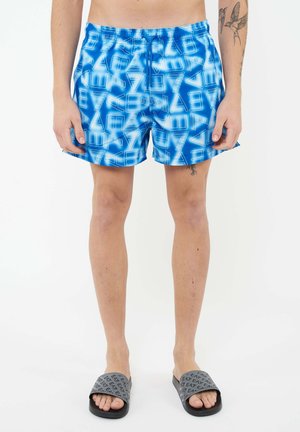 Uomo con pantaloncini da bagno blu a motivi e ciabatte grigie EA7, in piedi su sfondo bianco, con tatuaggi visibili su braccia e gambe.