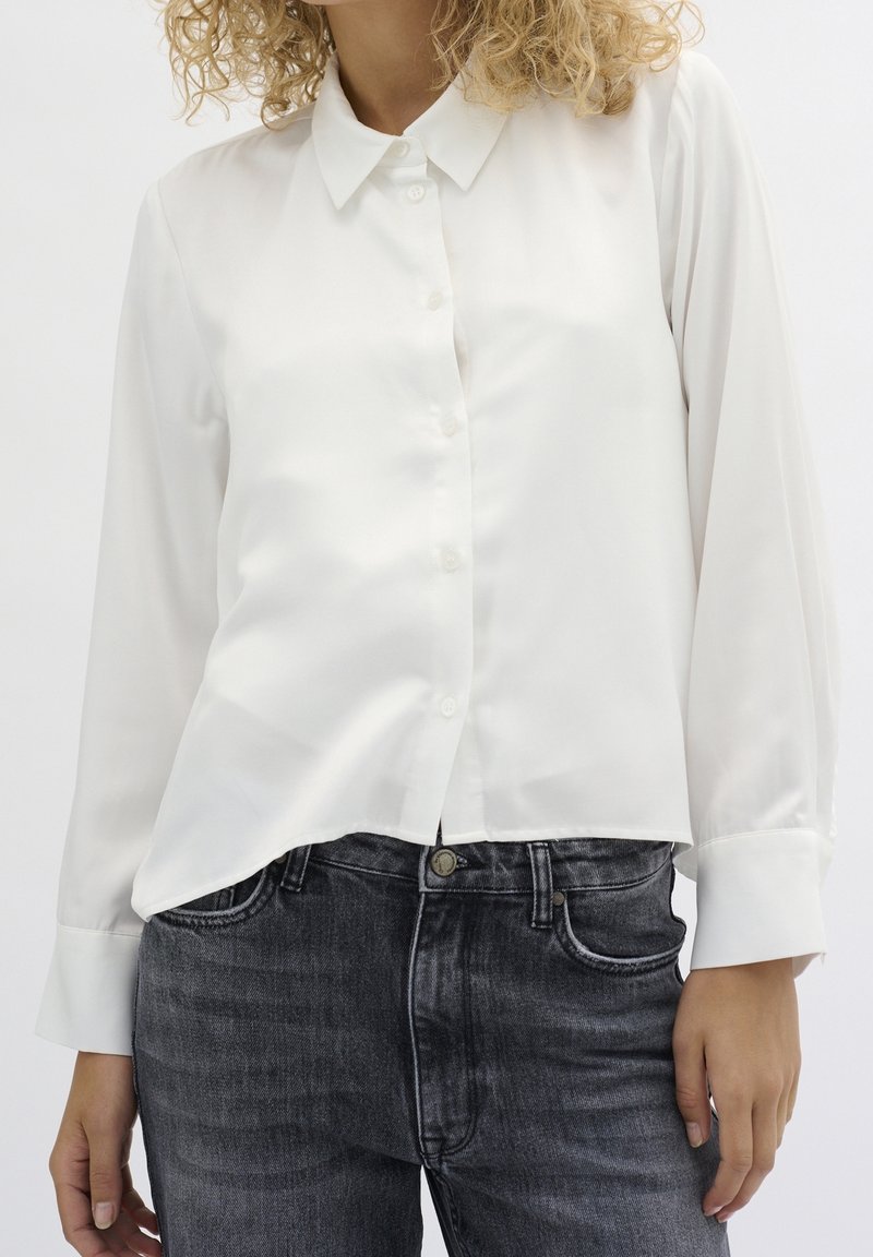 Femme portant une chemise blanche à manches longues, légèrement rentrée dans un jean noir délavé, debout avec les mains relâchées le long du corps.