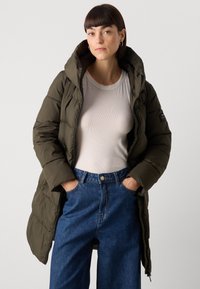 Olijfgroene pufferjas met een capuchon, geborduurde ontwerp, frontale rits en zijzakken. Draagt boven een witte fitted top en blauwe high-waisted jeans.