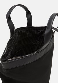 Sac fourre-tout noir avec une ouverture zippée, doté de poignées robustes et d'une doublure en tissu rayé. Le matériau est une combinaison de toile et de cuir.