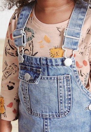 Małe dziecko ubrane w jeansowe ogrodniczki i beżową, długorękawową koszulkę z kolorowymi zwierzęcymi i abstrakcyjnymi wzorami.