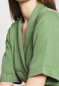 Top verde a maniche corte con scollatura incrociata, caratterizzato da una texture liscia e una vestibilità comoda, adatto per un abbigliamento informale.