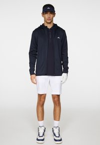 Completo sportivo da uomo: felpa con zip blu navy, polo scura, pantaloni corti bianchi, scarpe da ginnastica blu e bianche, guanto bianco, visiera blu navy, branding minimale.