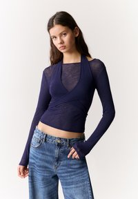 Dunkelblaues Langarmoberteil mit Wickeldesign, aus leichtem Stoff gefertigt. Verfügt über einen hohen Hals und Daumenlöcher. Vorgesehen in Kombination mit hochtaillierter Jeans.