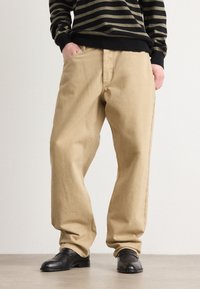 Pantalon beige clair à coupe décontractée, fabriqué dans un tissu texturé. Présente des poches avant et des coutures contrastées, associé à des chaussures noires.