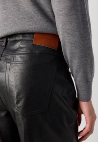 Černé kožené kalhoty mají hladkou strukturu a viditelné švy. Na zadní straně je kožená nášivka v odstínu tan s nápisem "Calvin Klein". Šedý svetr je částečně viditelný.