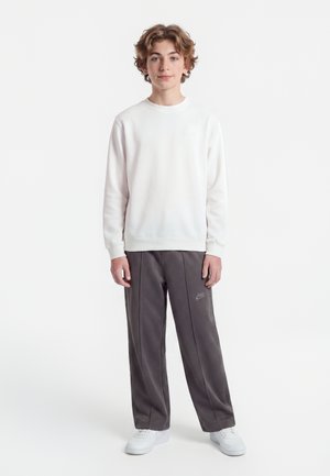 Teenage dreng stående iført hvid sweatshirt, mørkegrå Nike træningsbukser og hvide sneakers mod en ensartet lys baggrund.