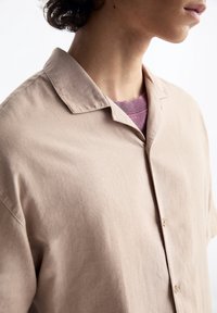Camisa de botones de manga corta en color beige, confeccionada en una tela ligera con una textura suave. Presenta un collar con muesca y un color neutro sutil.