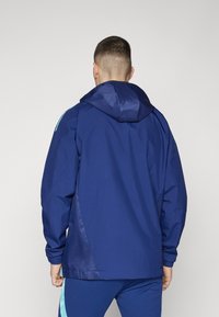 Giacca con cappuccio blu navy dal tessuto liscio, con polsini elasticizzati e dettagli a contrasto blu chiaro sulle spalle. Leggera e dalla vestibilità oversize.