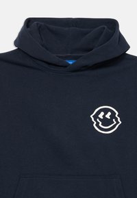 Sudadera con capucha de color azul marino hecha de un tejido suave, presenta un logo de cara sonriente blanca en el lado izquierdo del pecho y un bolsillo tipo canguro en la parte frontal.