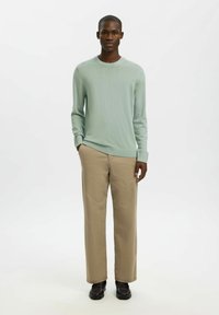 Pull en maille vert clair avec un col rond, associé à un pantalon beige. Le pantalon a une coupe droite et est porté avec des chaussures foncées.