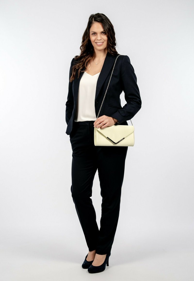 Marinblazer med matchande byxor, vit blus och svarta högklackade skor. Bär en gul kuvertväska med kedjerem.