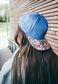 Blue baseball cap with a floral-patterned brim and a brown leather tag accent. Smooth texture, worn tilted back, revealing long hair.