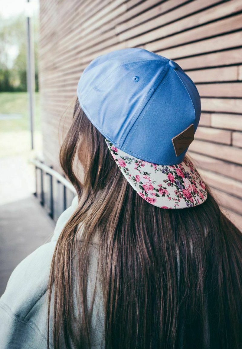 Blue baseball cap with a floral-patterned brim and a brown leather tag accent. Smooth texture, worn tilted back, revealing long hair.