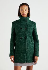 Manteau en laine texturée verte avec un col haut, des boutons noirs doubles et des poches latérales, présentant une coupe droite et une coupe décontractée.