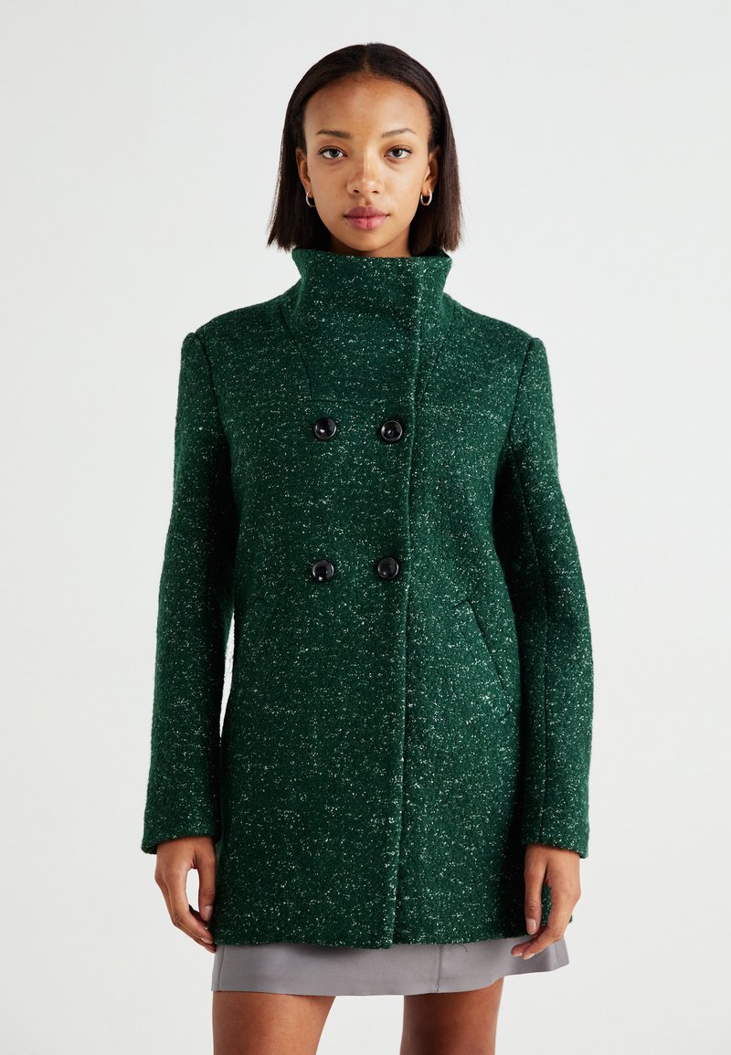 Manteau en laine texturée verte avec un col haut, des boutons noirs doubles et des poches latérales, présentant une coupe droite et une coupe décontractée.