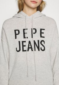 Femme portant un sweat à capuche gris clair avec le texte noir "PEPE JEANS" sur la poitrine, debout devant un fond uni.