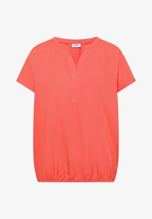 Femme aux cheveux courts bruns portant un chemisier corail à manches courtes avec un pli dans le dos et un jean bleu clair, tournée légèrement de côté.
