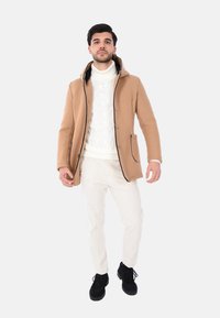 Cappotto con cappuccio color cammello con interno nero, maglione bianco lavorato a trecce, pantaloni in velluto a coste color crema e stivali neri. Taglio slim, tasche laterali visibili.