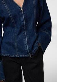 Tumesinistest vertikaalsete õmblustega ja laia varrukaga denimjakk esiküljel lukuga. Kleiditud mustade teksadega, väljendades vabaaja stiili.