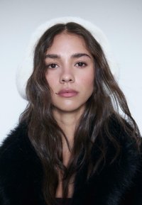 Sorte pelsjakke og hvidt faux pels hat. Lang, bølget mørkt hår. Naturlig makeup med et koncentreret udtryk. Huden ser glat ud. Neutral baggrund.