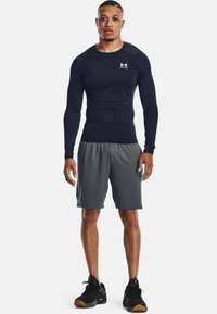 Langermet marinblå treningsskjorte kombinert med grå shorts. Skjorten har en tettsittende design; shortsene har et strukturert mønster. Svarte treningssko fullfører antrekket.