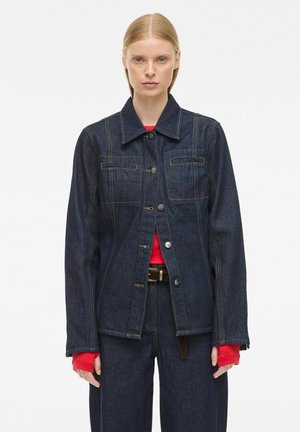 Vrouw in een donkerdenim jack en broek met daaronder een rood shirt met lange mouwen, staande tegen een effen witte achtergrond.