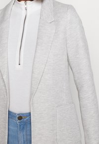 Světle šedý blazer s herringbone vzorem, s jednou kapsou a ostrými klopami, nošený přes bílý top s vysokým krkem a světle modré džíny.