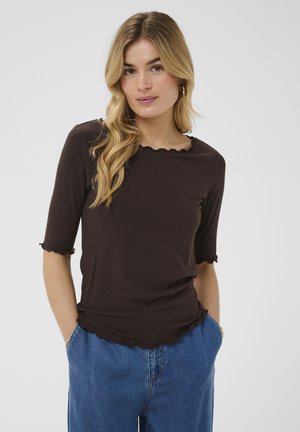 Femme blonde aux cheveux ondulés porte un haut noir côtelé avec des bords festonnés et un pantalon en jean bleu, mains dans les poches, arrière-plan neutre.
